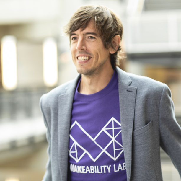 Jon Froehlich is a white man with brown hair wearing a purple Makeability Lab t-shirt and a gray sports coat