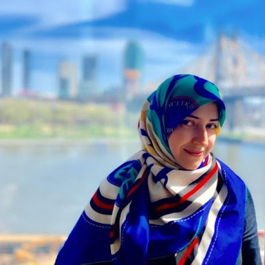 Maryam Hosseini is standing in front of a beautiful urban scene with a river and skyline. She has brown hair and eyes and is wearing a hijab.