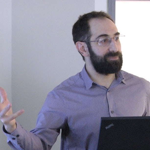 Yochai Eisenberg is standing in front of a podium giving a talk. He has brown hair, a beard, and is wearing a purple shirt